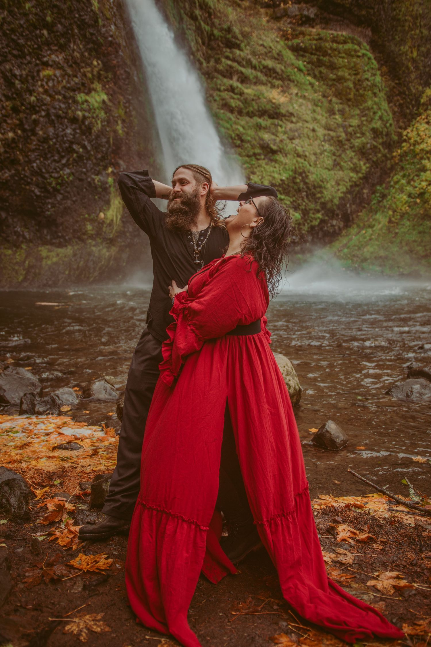 Couple strikes dramatic pose against waterfall backdrop with red dress flowing in the breeze.