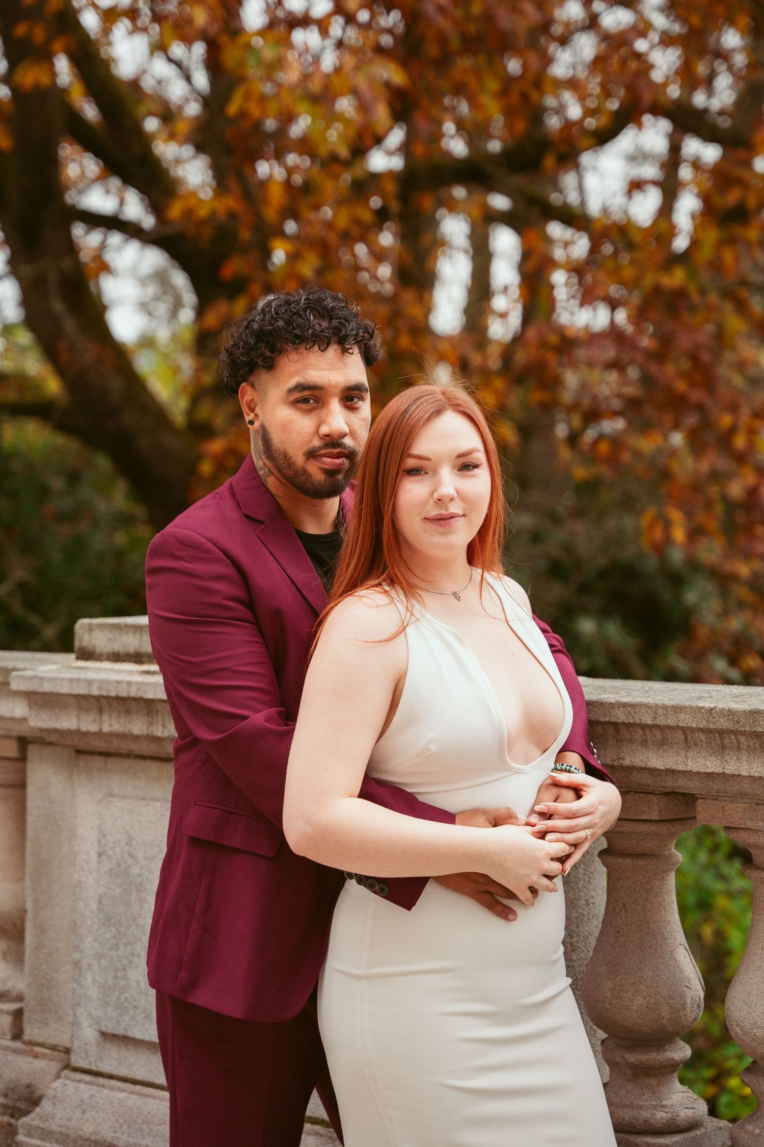 A couple embraces against an autumn backdrop with orange foliage and stone balustrades.