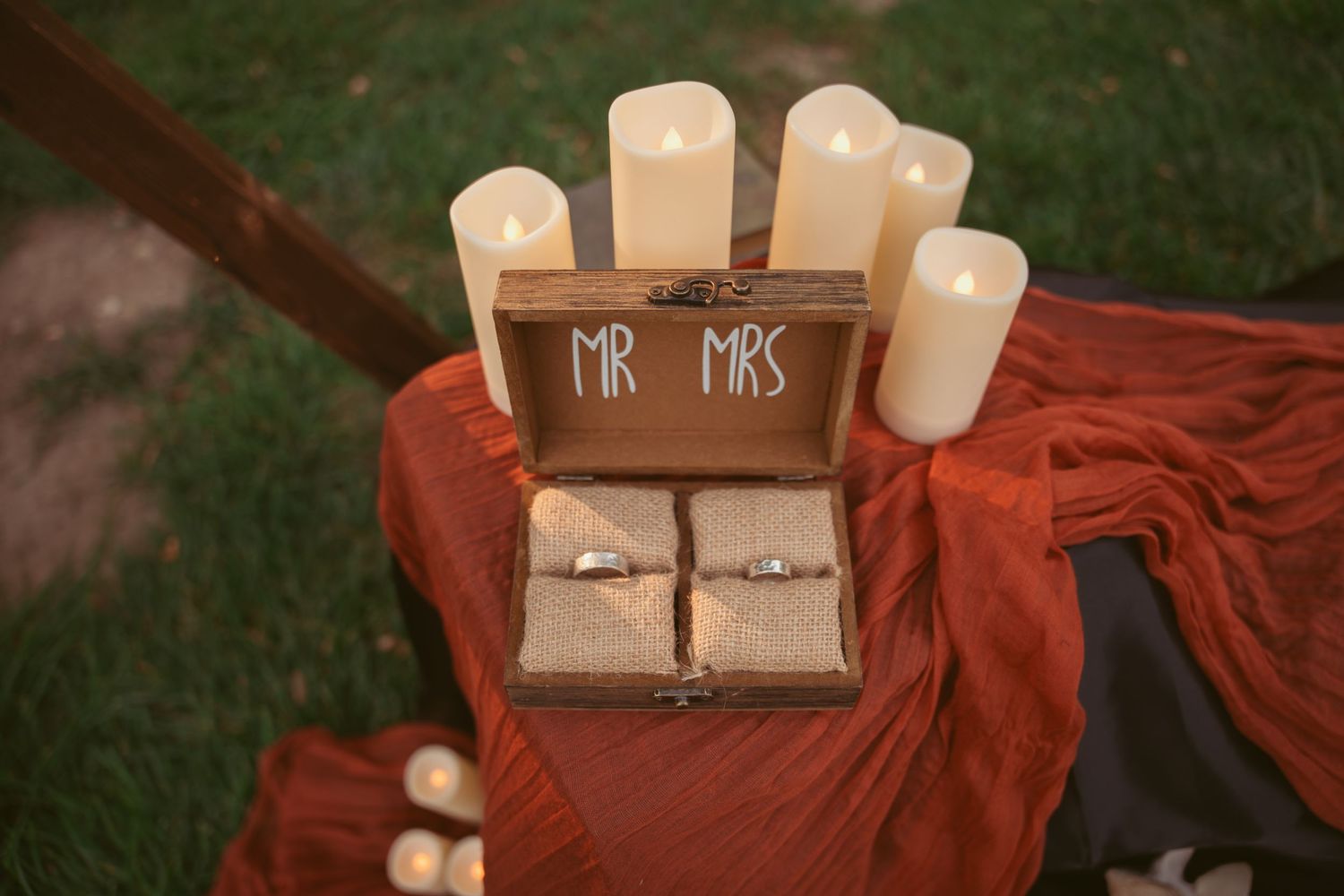 White candles and wooden decor elements arranged on a red tablecloth for a rustic outdoor wedding setup.