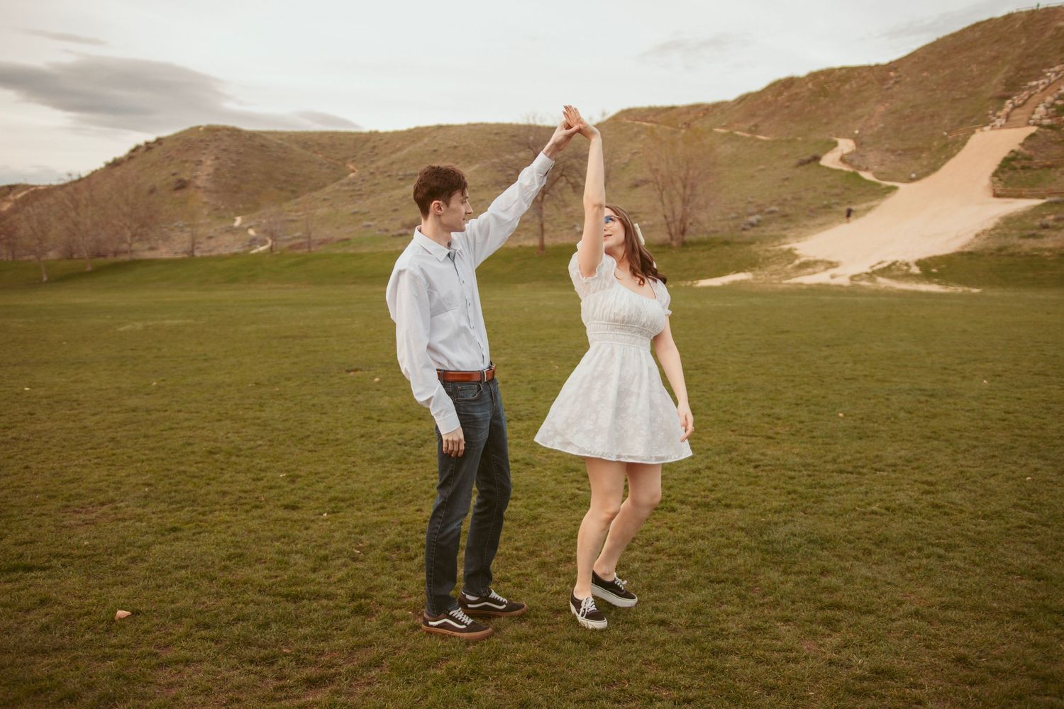 Couple dancing and twirling in white outfits on grassy hillside.