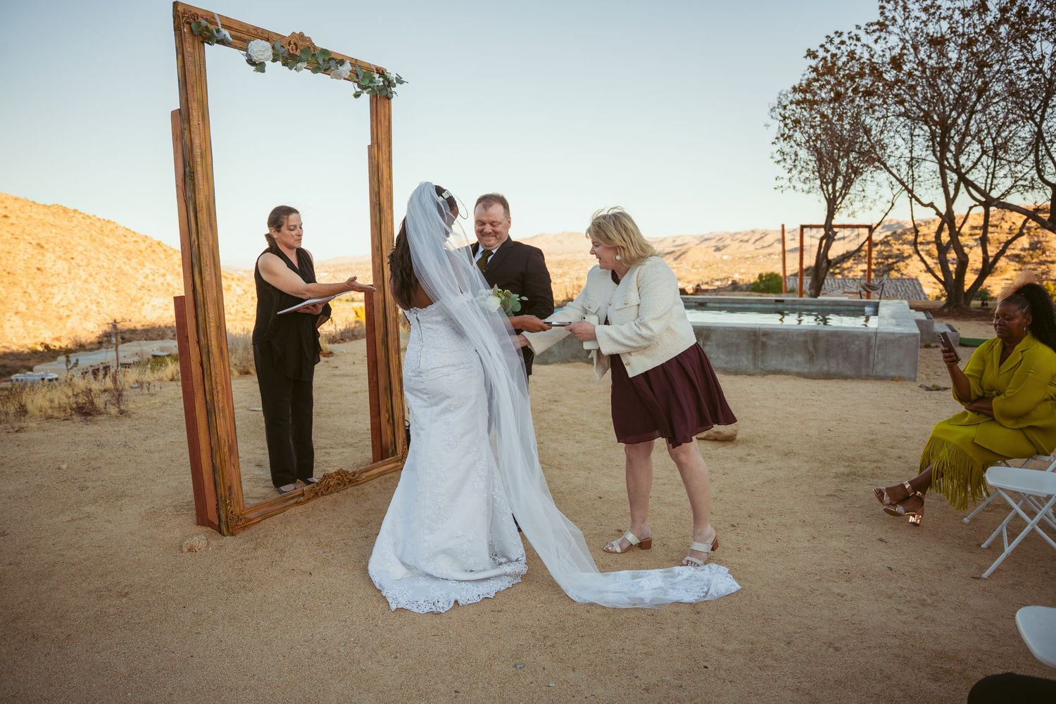 Desert wedding ceremony with wooden arch and intimate gathering of guests.