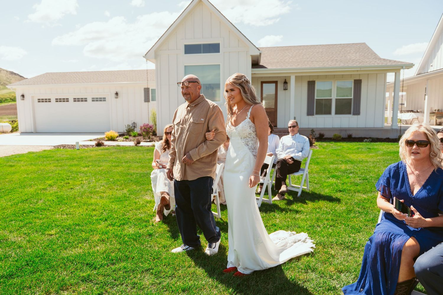 White farmhouse style buildings and wedding guests gathered on a sunny lawn for an outdoor celebration.