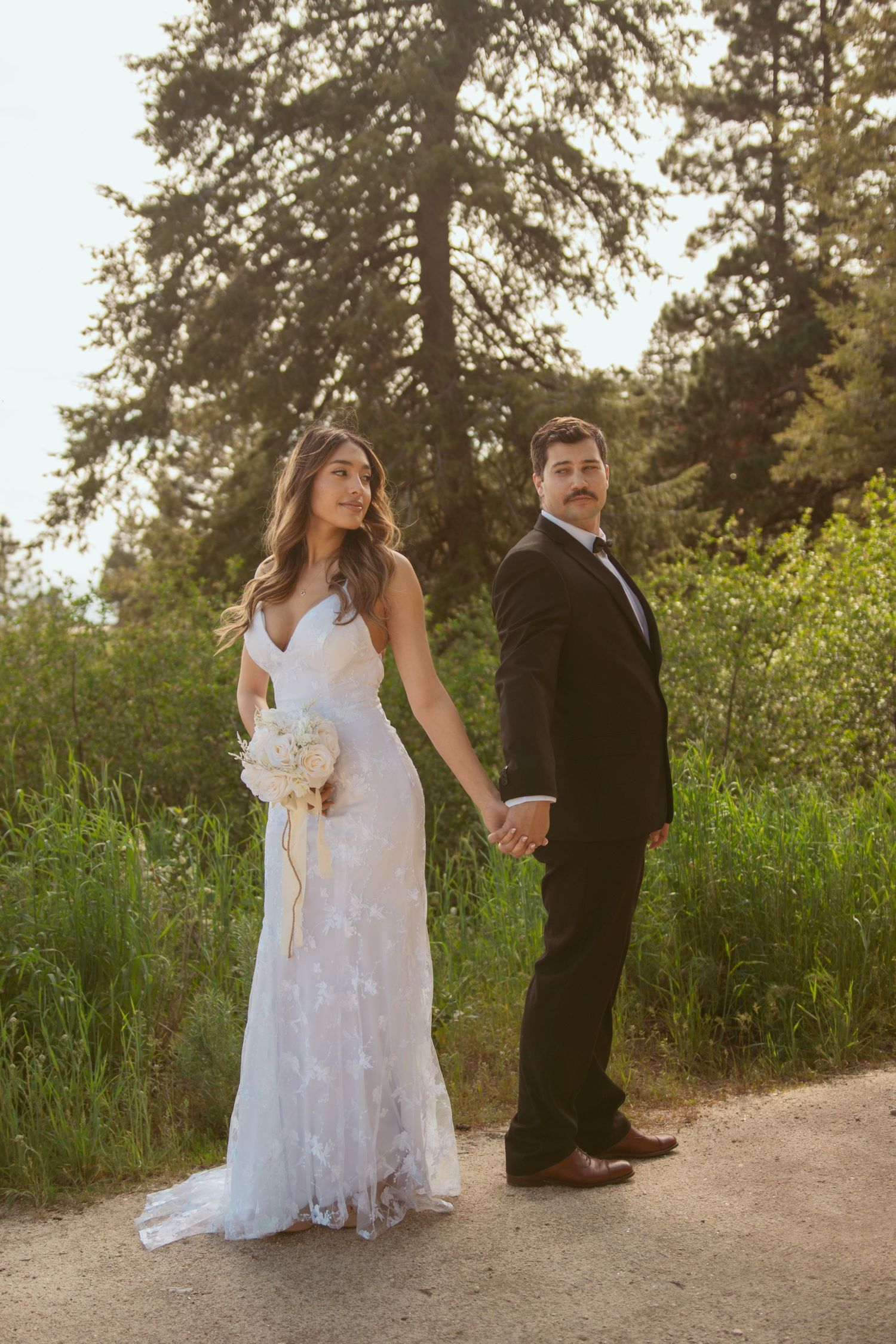 A couple in wedding attire holds hands while standing on a dirt path surrounded by evergreen trees at sunset.