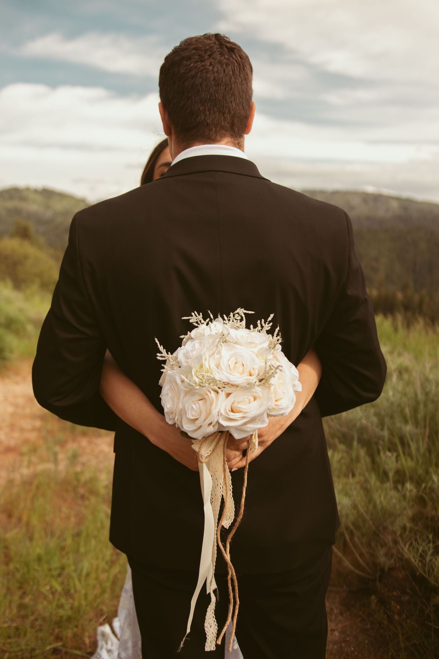 Back view of a person in a black suit holding a white rose bouquet against a mountain landscape.