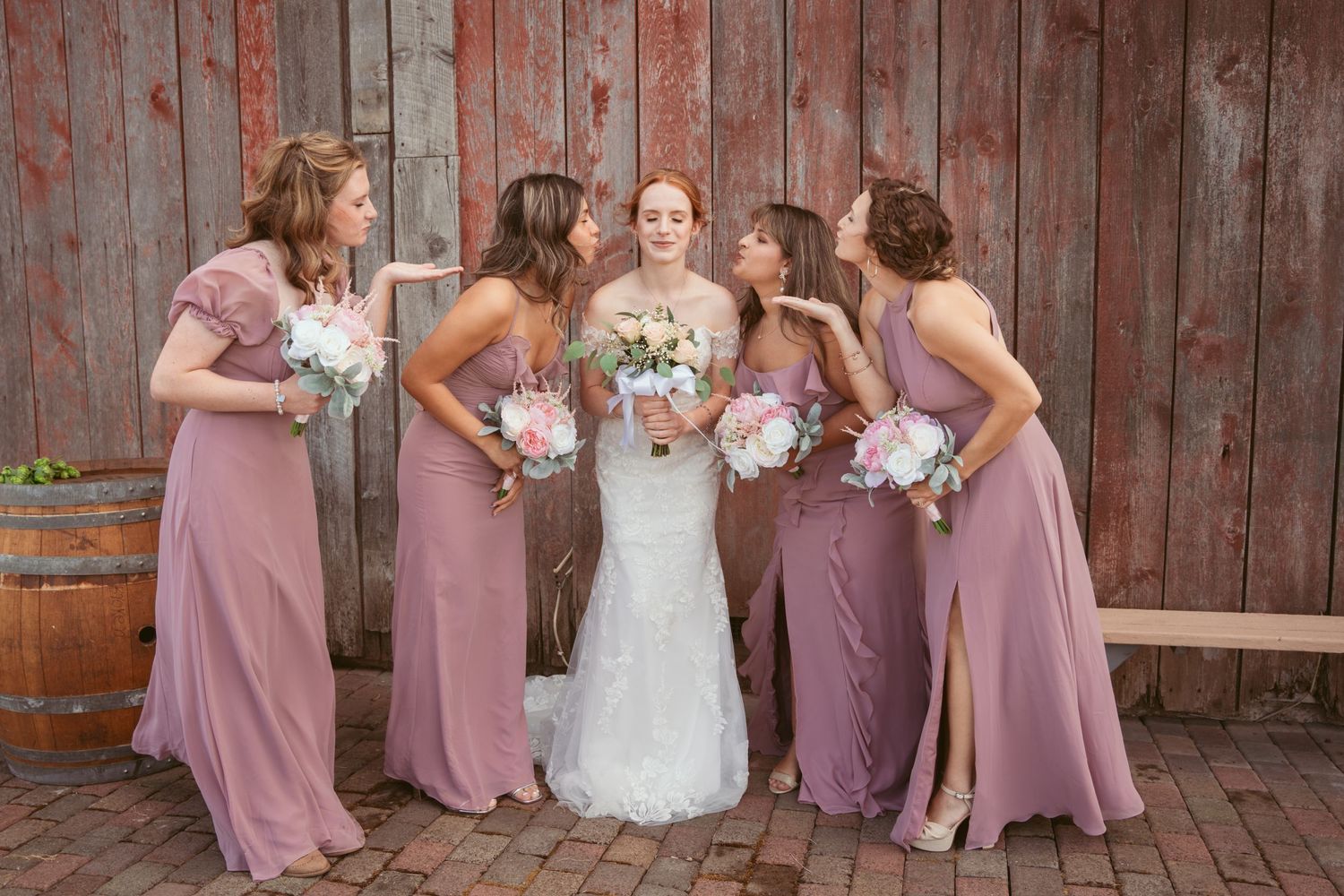 Wedding party members in mauve dresses and white bridal gown pose together by rustic barn.