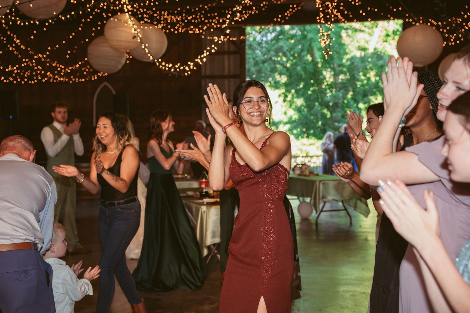 Wedding party dancing and celebrating under twinkling lights and lanterns at a rustic indoor venue.