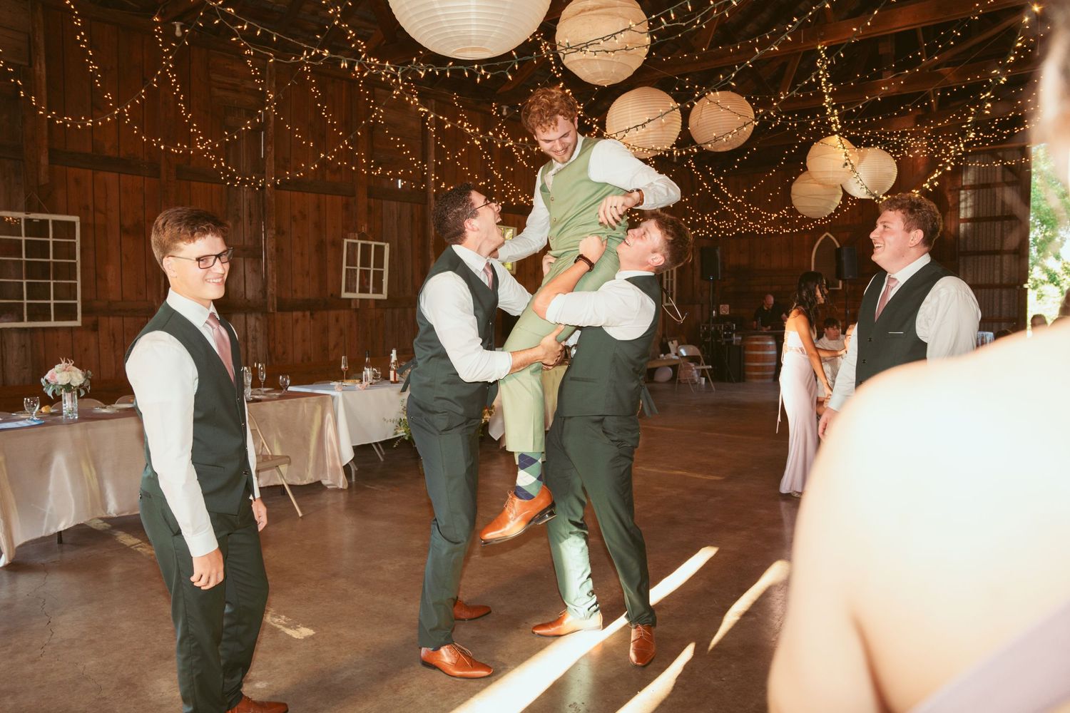 Wedding party dancing in coordinated outfits under string lights in a rustic barn setting.
