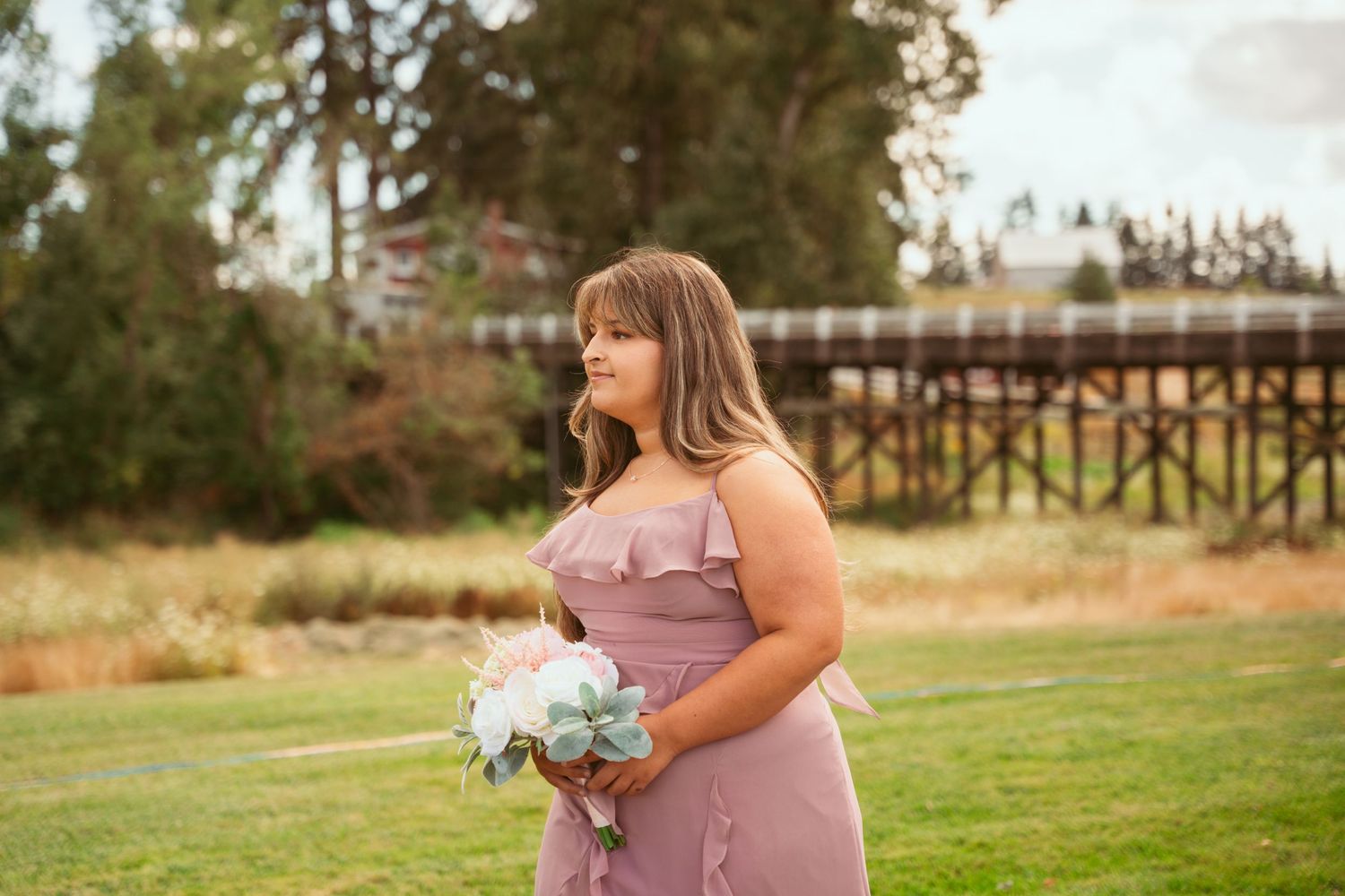 A person in a dusty rose dress carries a bouquet while walking on a brick path at an outdoor event.