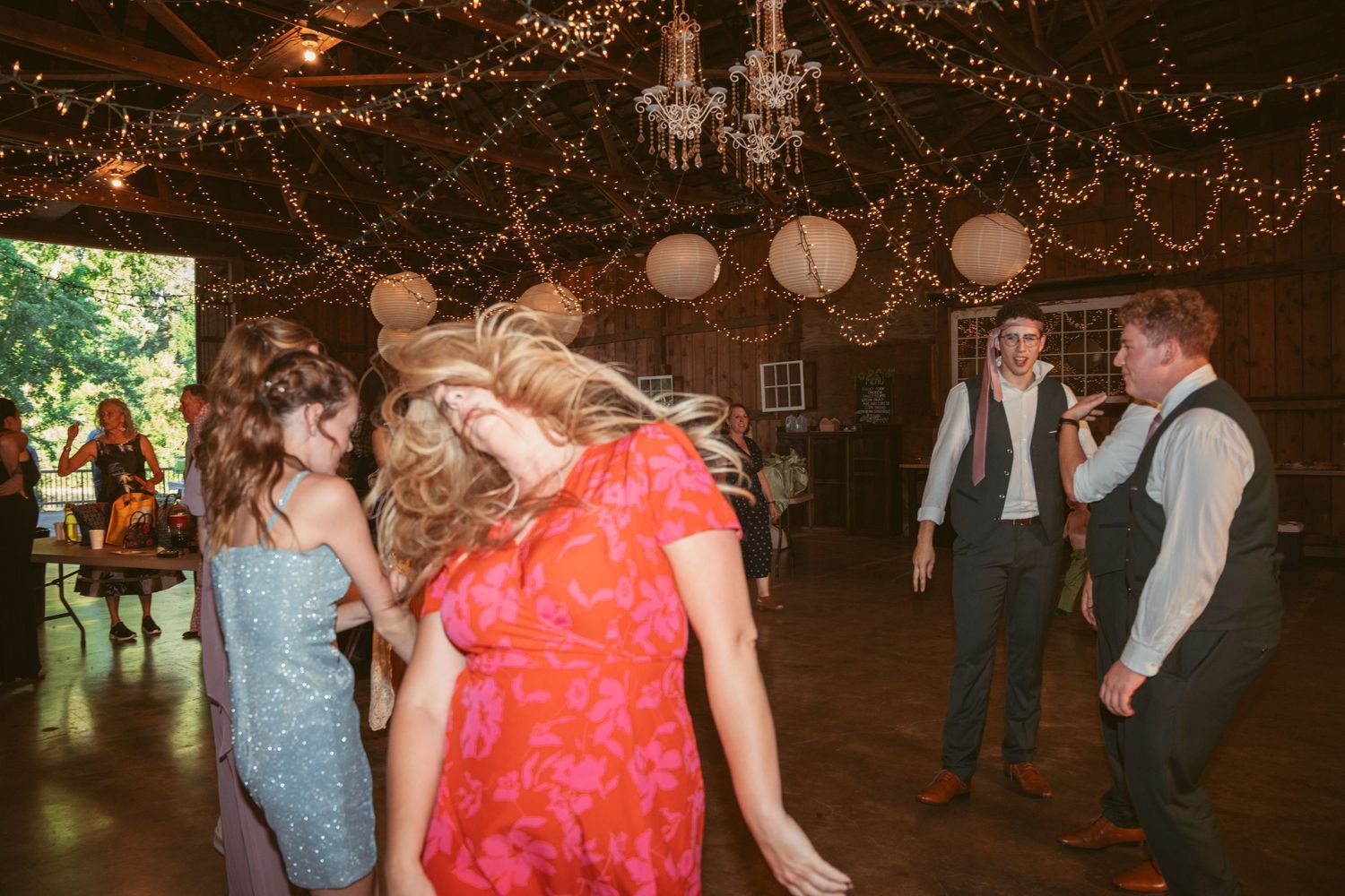 Party guests dancing under string lights and paper lanterns in a rustic barn setting.