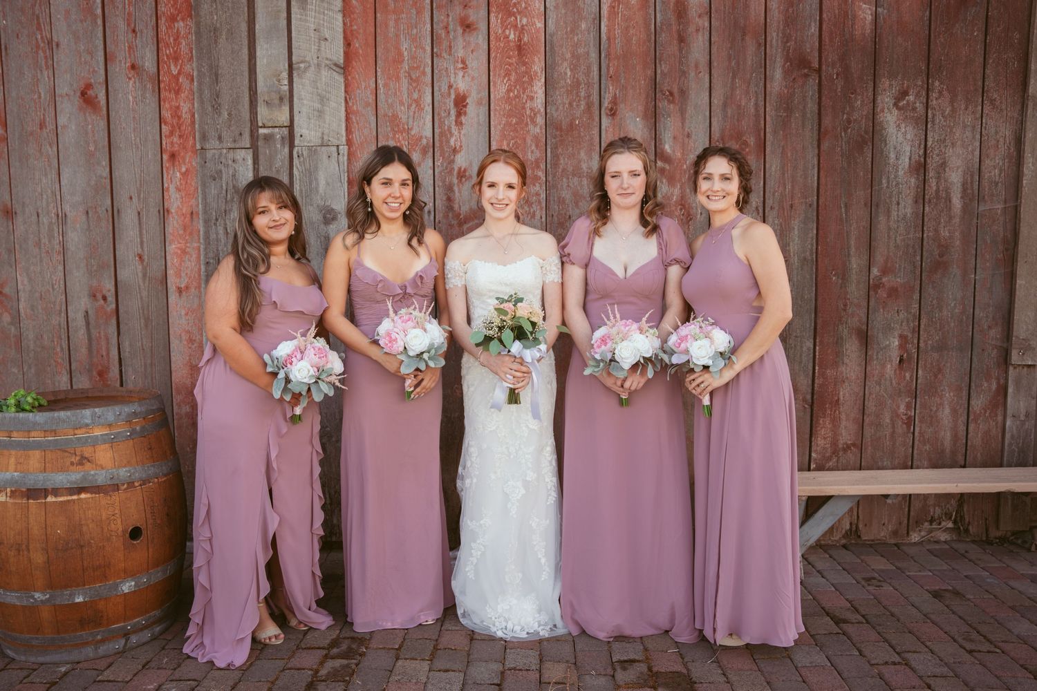 Bridal party in mauve dresses and white wedding gown pose against rustic red barn backdrop.