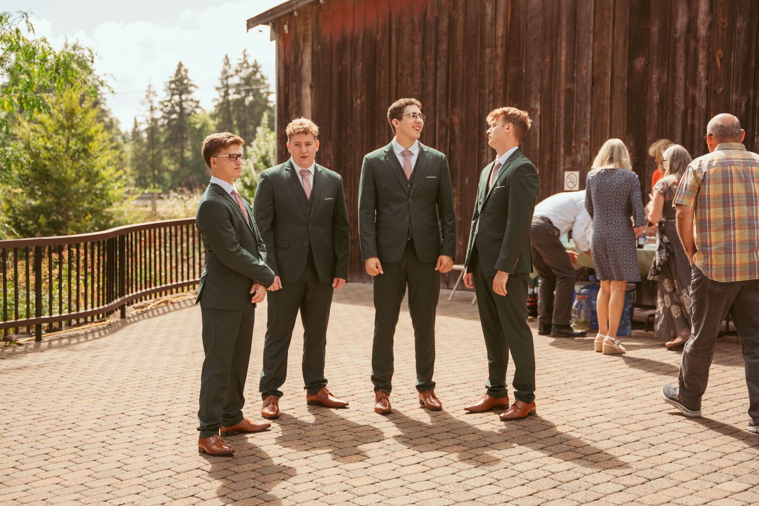 Groomsmen in dark suits gather on a brick patio outside a rustic barn venue.