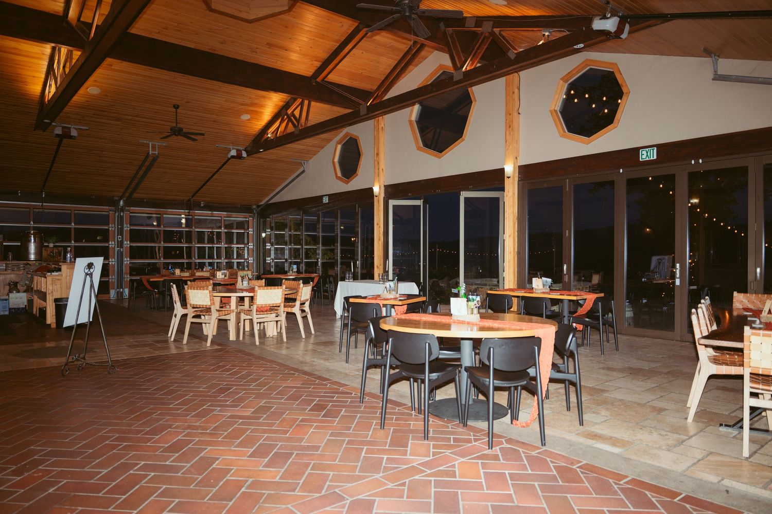 Large indoor reception venue with exposed beams, brick flooring, and scattered round dining tables.