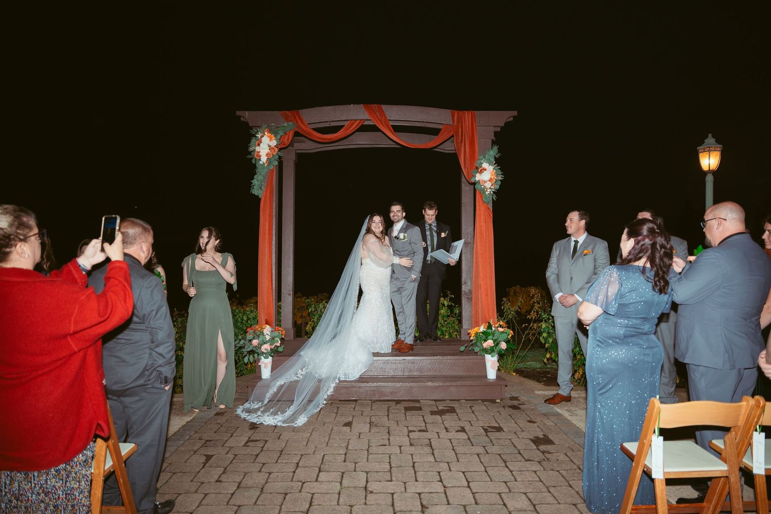 Wedding celebration under string lights with wooden chairs arranged on stone patio.
