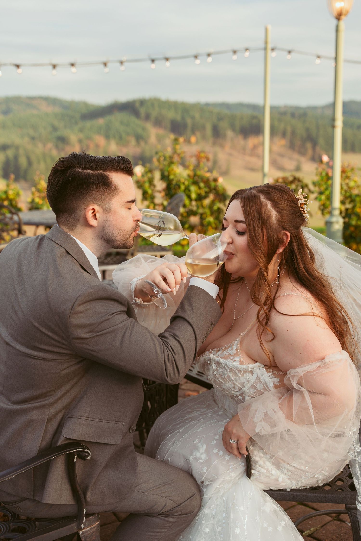 Wedding couple shares playful wine drinking moment at outdoor vineyard reception.
