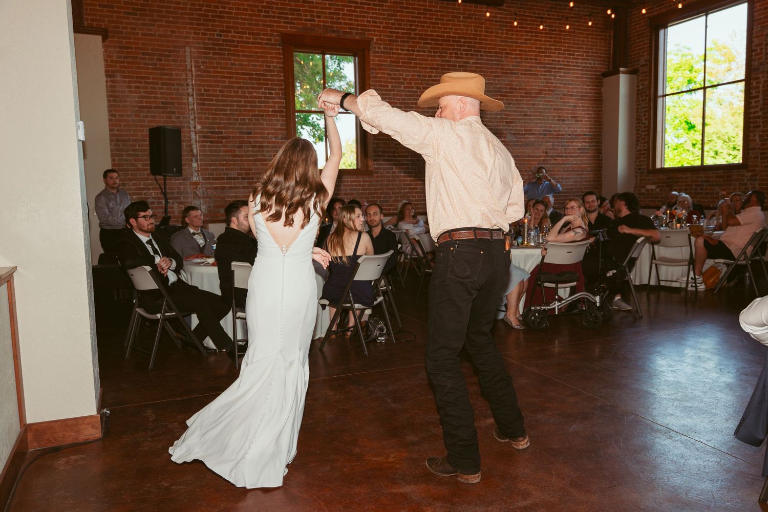Wedding couple performs a dramatic dance dip move in a rustic brick venue with string lights overhead.