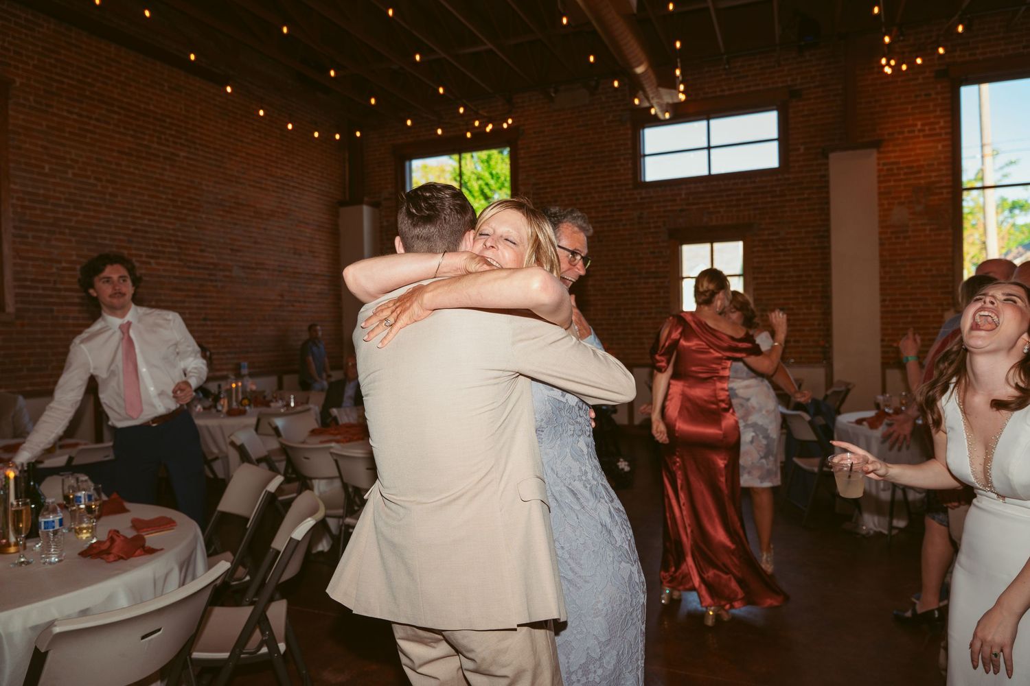 Guests dance together at a wedding reception in an industrial-style venue with string lights.