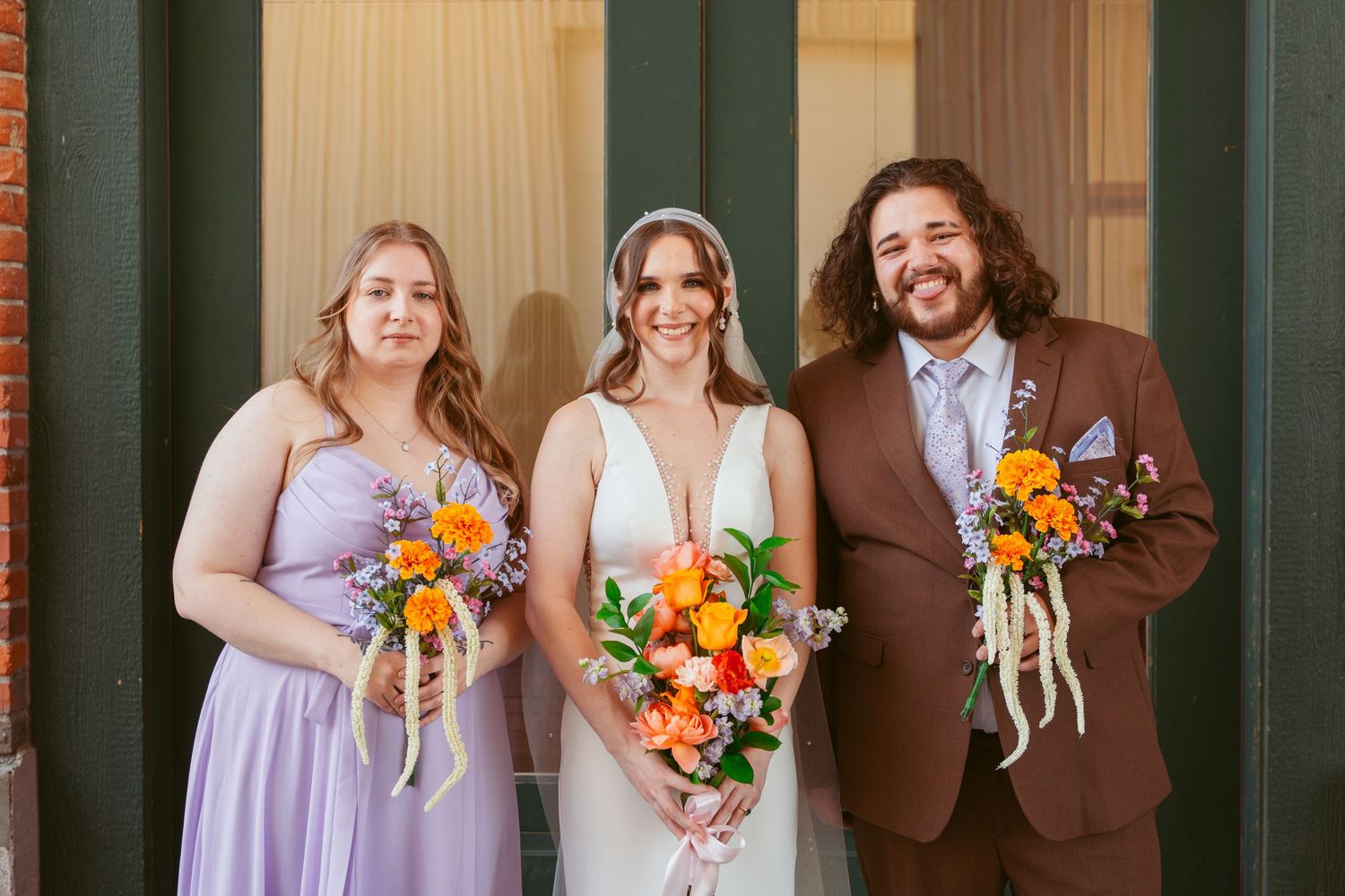 Wedding party of three poses with bright orange and pink floral bouquets against green doorway.