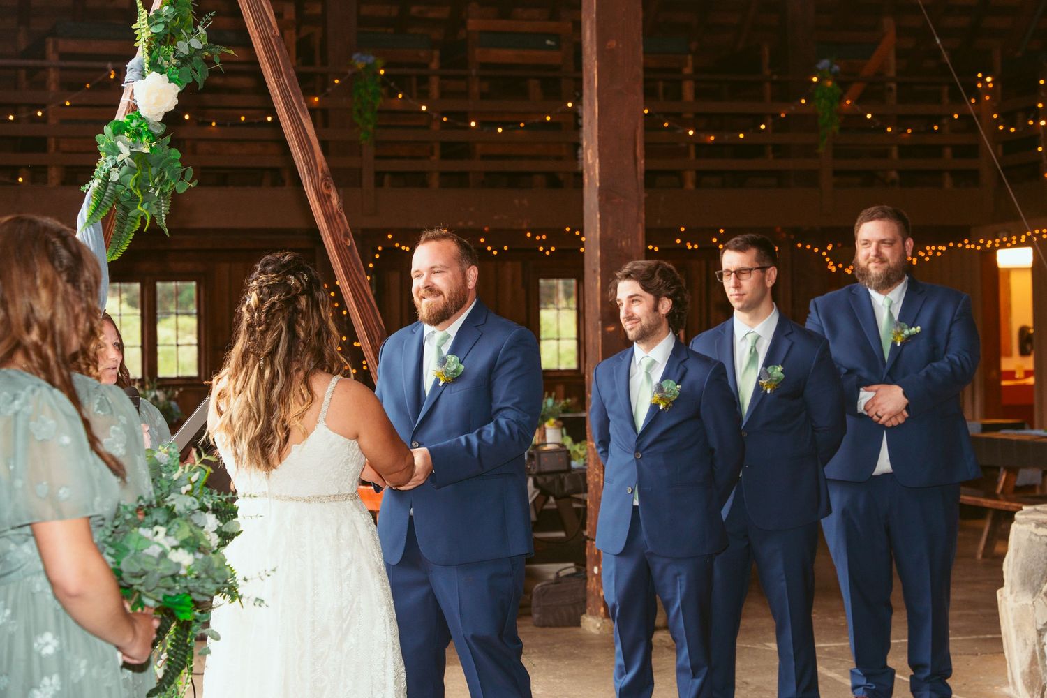 Wedding party lined up in navy attire inside rustic barn with string light decorations.