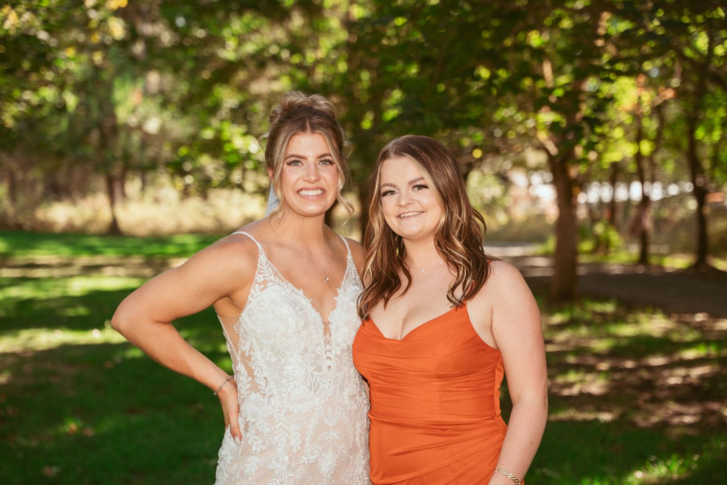 Two women in white and orange dresses pose together in a sunny outdoor garden setting.