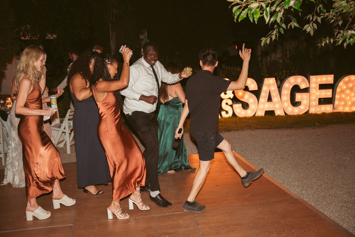 Wedding guests dancing in formal attire near illuminated signage at outdoor reception.