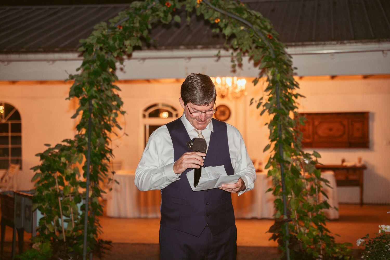 A speaker reads from papers under an illuminated vine-covered arch during an evening event.