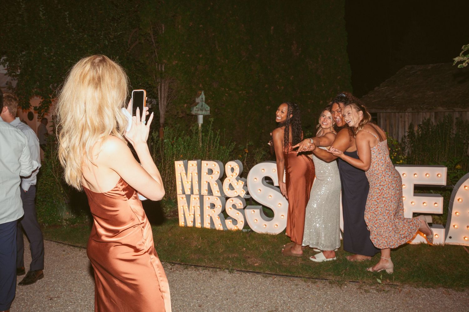 Wedding guests dancing and celebrating at an outdoor evening reception with illuminated MRS sign in background.
