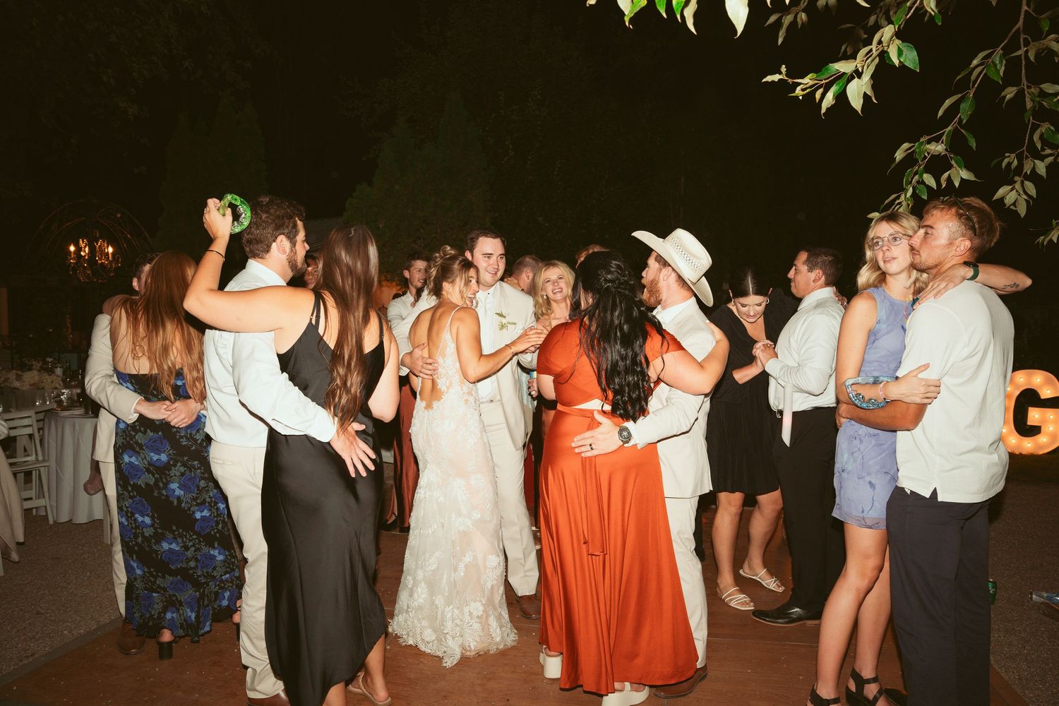 Wedding party and guests dancing together at outdoor evening reception.