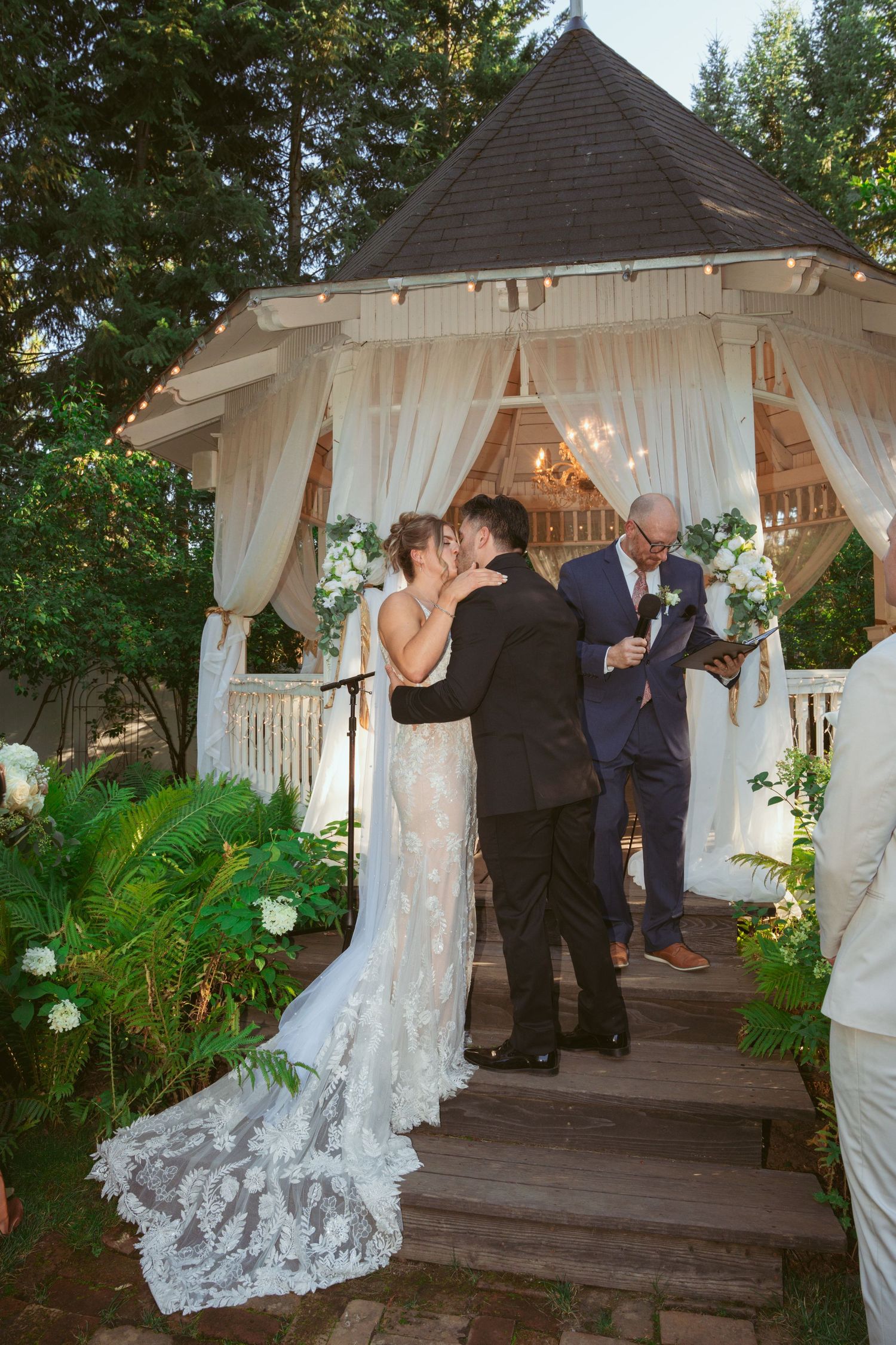 Wedding couple shares their first kiss under a decorated gazebo with white drapes and floral arrangements.