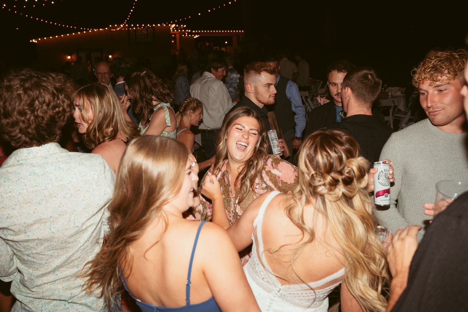 A lively group of party guests dancing and celebrating on a crowded dance floor under string lights.