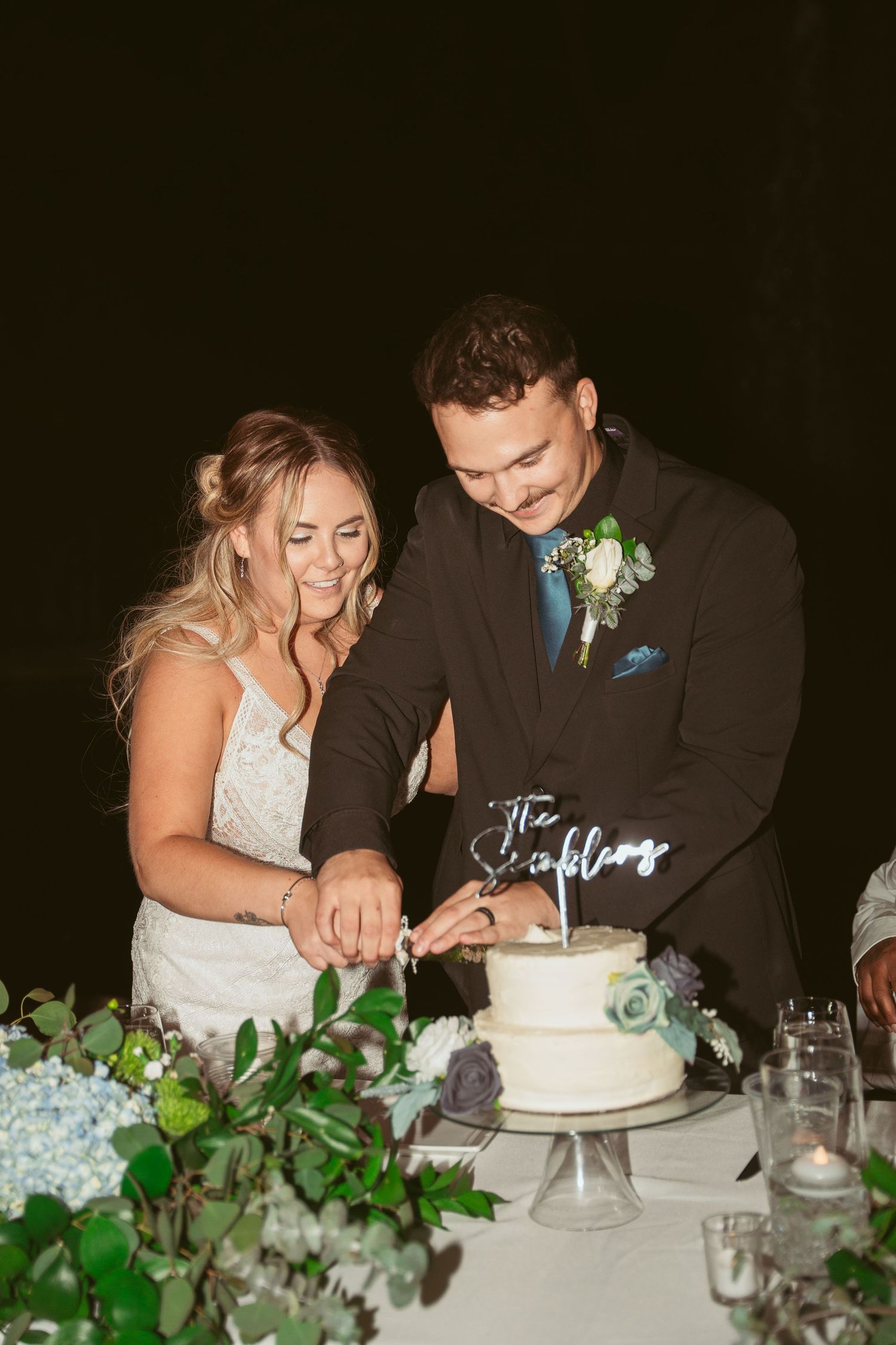 Newlyweds cut their white wedding cake together at the reception decorated with greenery.