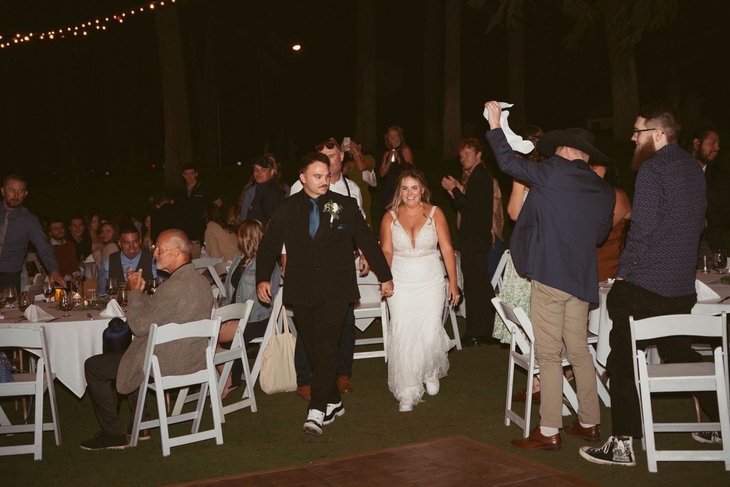 Wedding reception guests seated at tables with white chairs under string lights at night.
