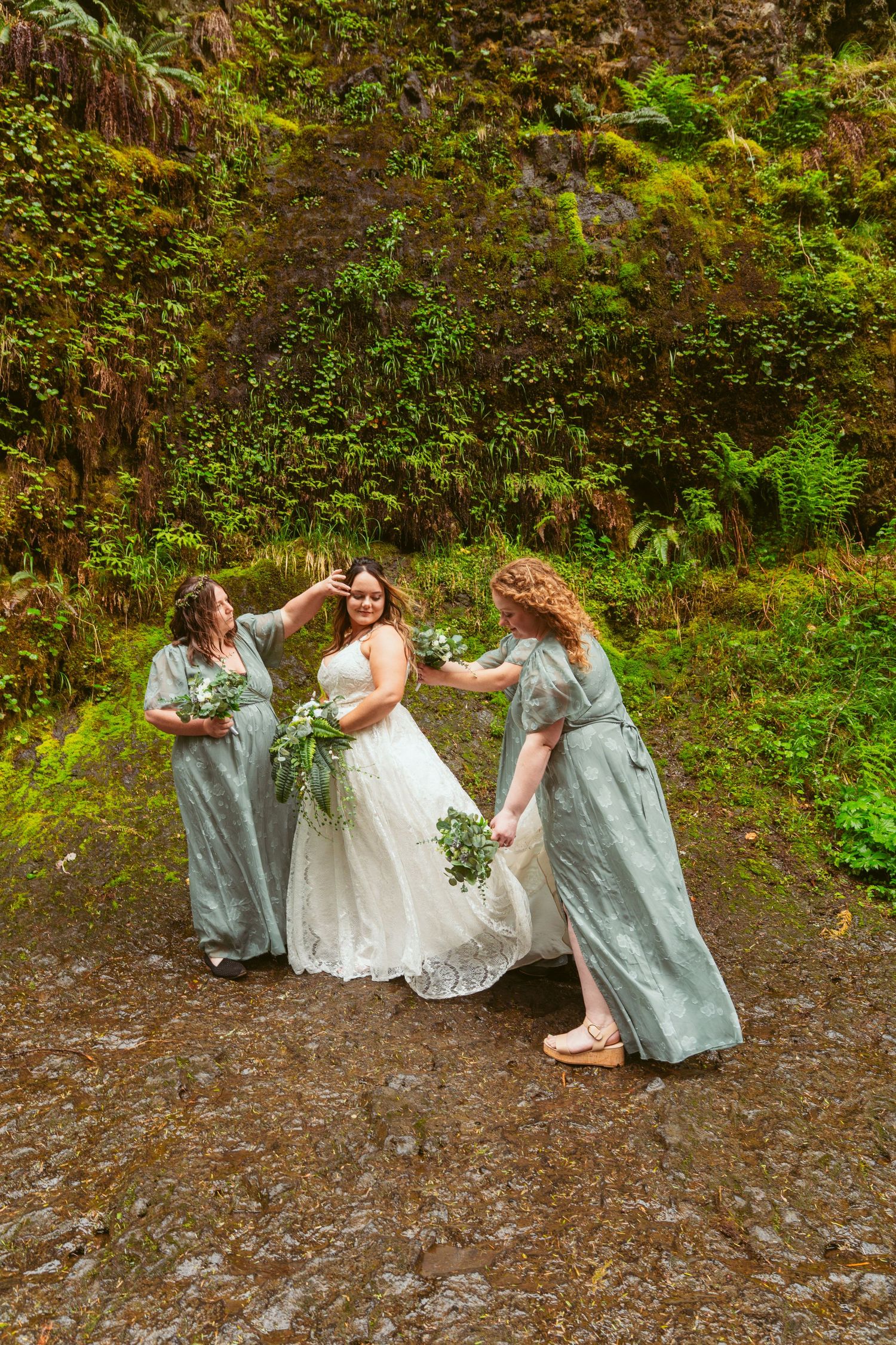 Bridesmaids assist with arranging the bride's dress during an outdoor photo session.