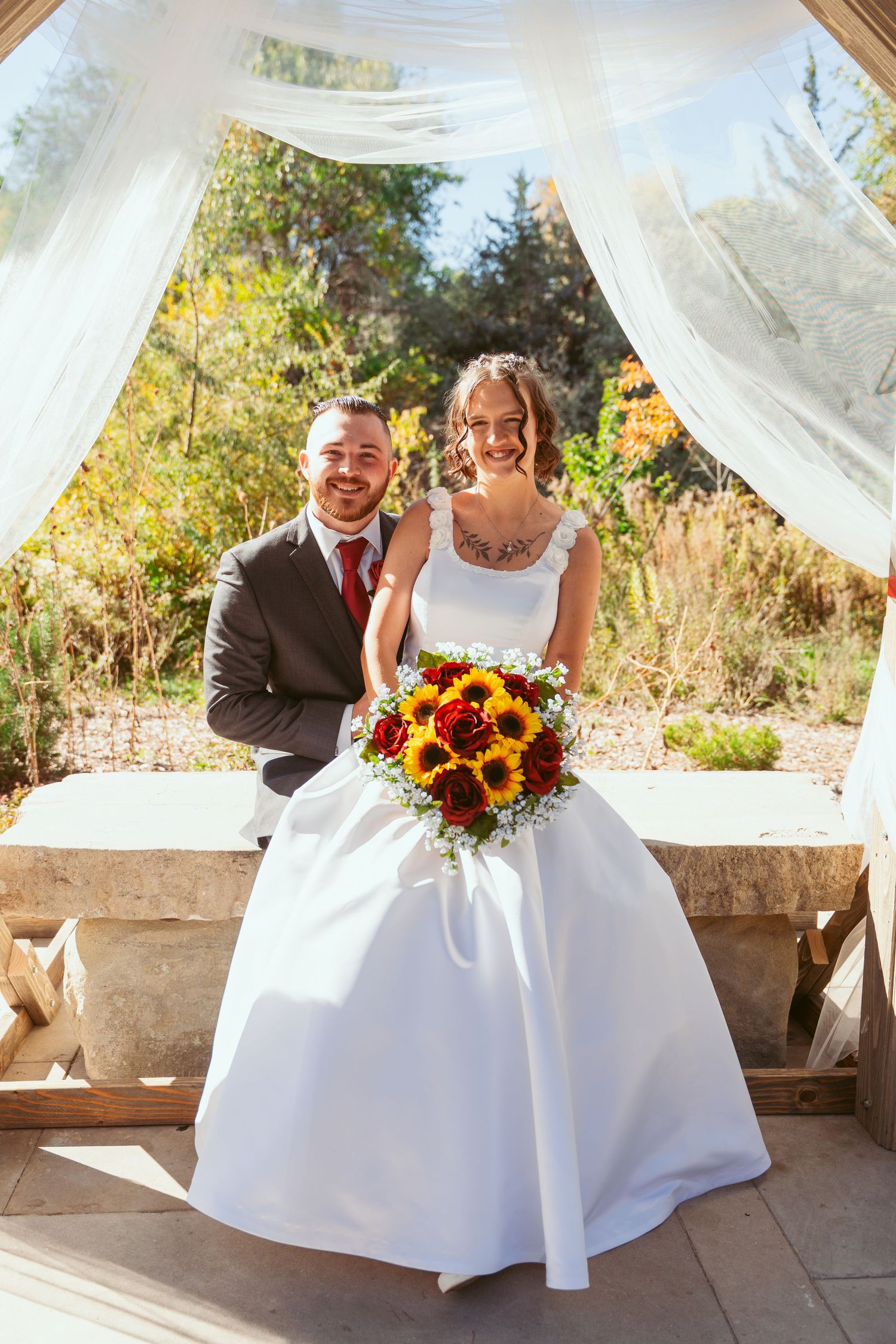 Wedding couple poses playfully on stone bench under draped pavilion with sunflower bouquet.