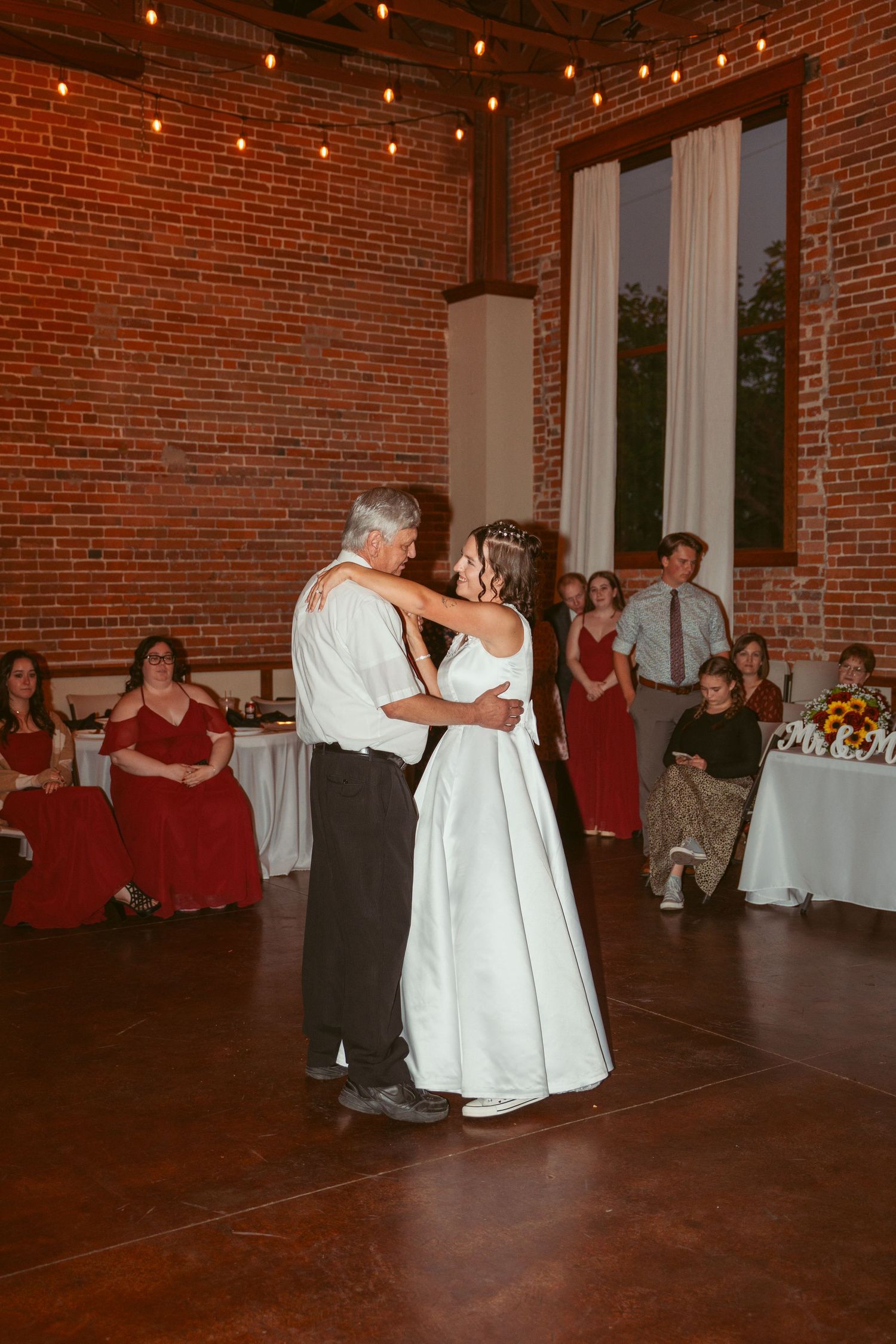 A couple dances at a wedding reception in a rustic brick venue with string lights overhead.