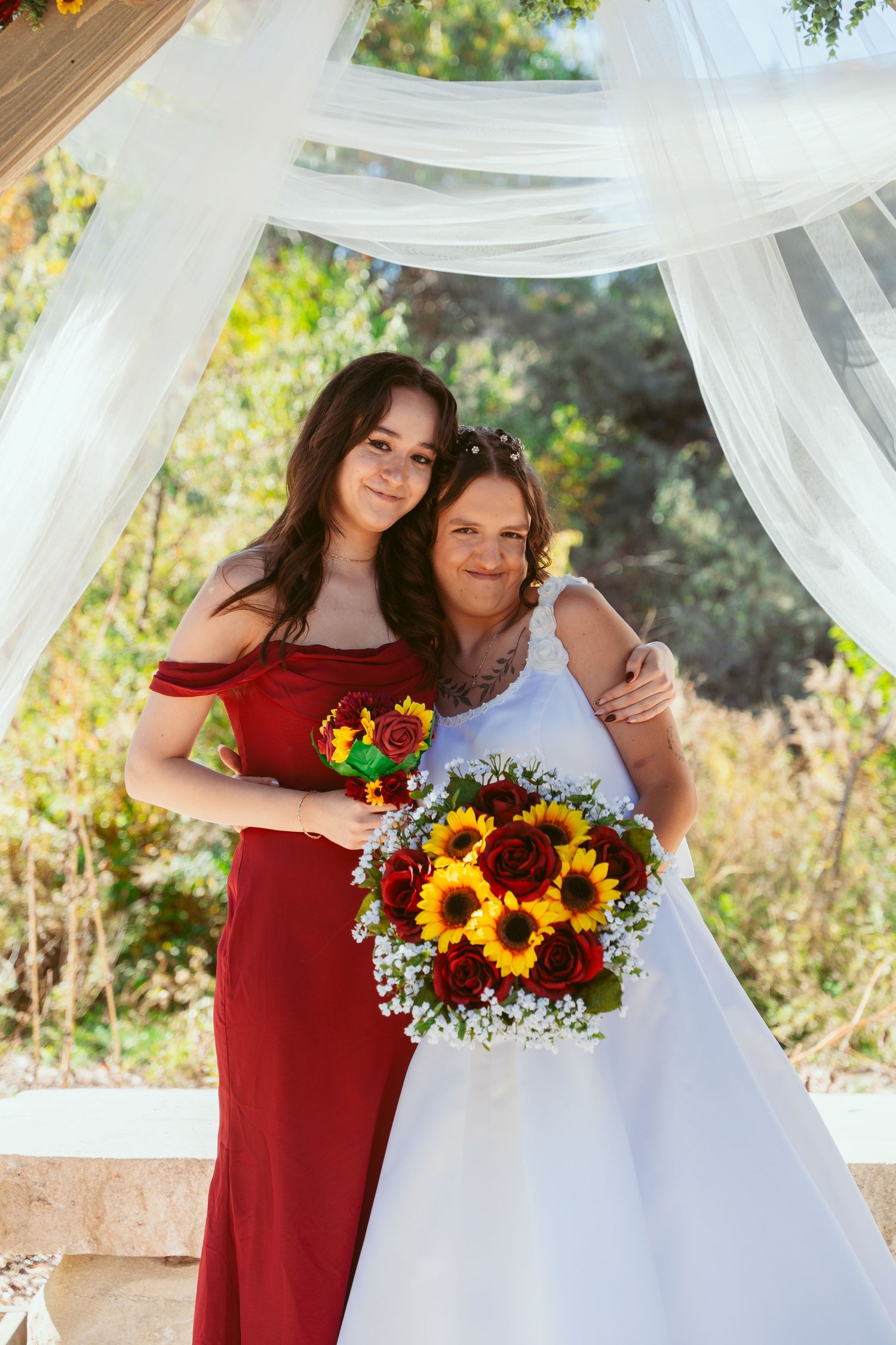 Wedding party members in red and white dresses pose together under a decorated outdoor pavilion with sunflowers.