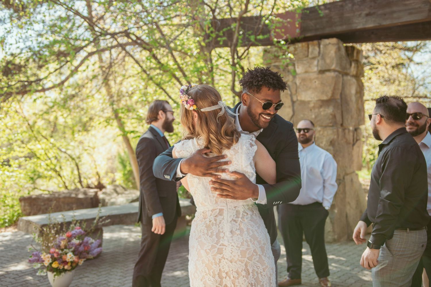 A romantic outdoor wedding scene shows a couple embracing under a rustic wooden pergola during their ceremony.