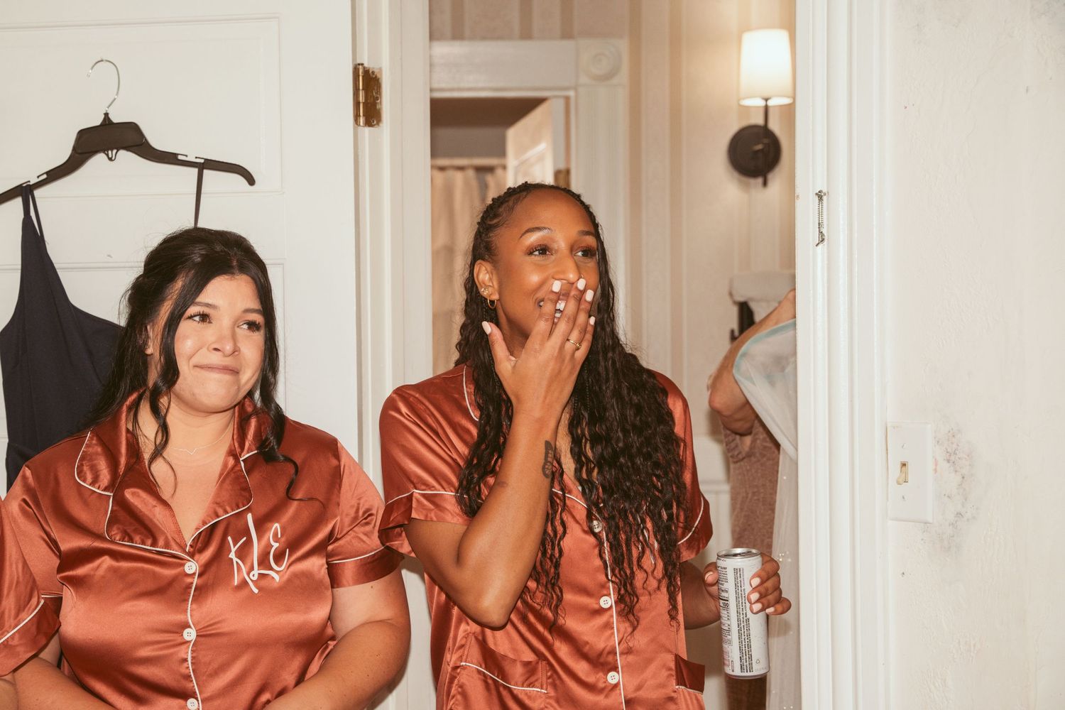 Two bridesmaids in matching silk pajamas react with surprise during wedding preparations.