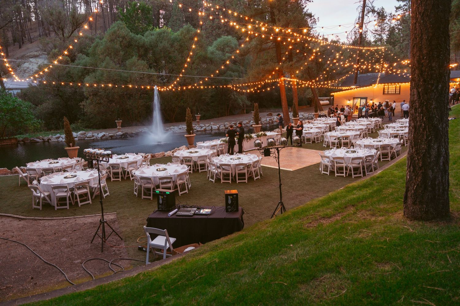 String lights illuminate an outdoor wedding reception venue with round tables set up under tall trees at dusk.