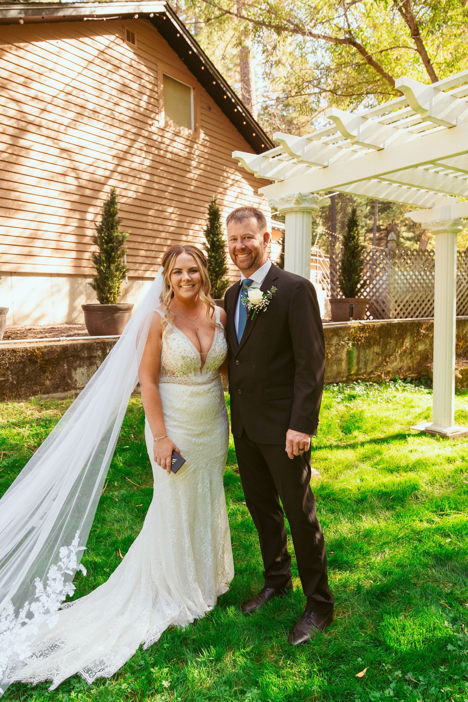 Bride in flowing veil and groom share candid moment in sunlit garden setting.