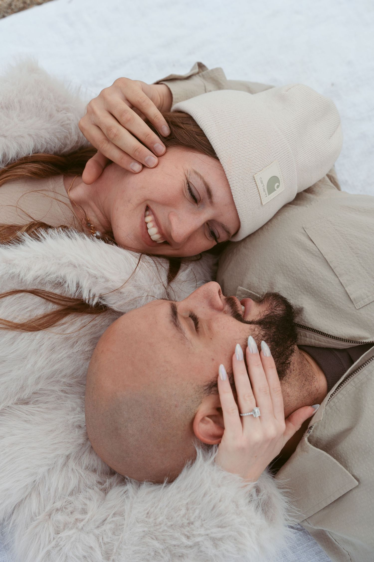 A couple lying on a white fluffy rug laughing together while wearing matching beige outfits.