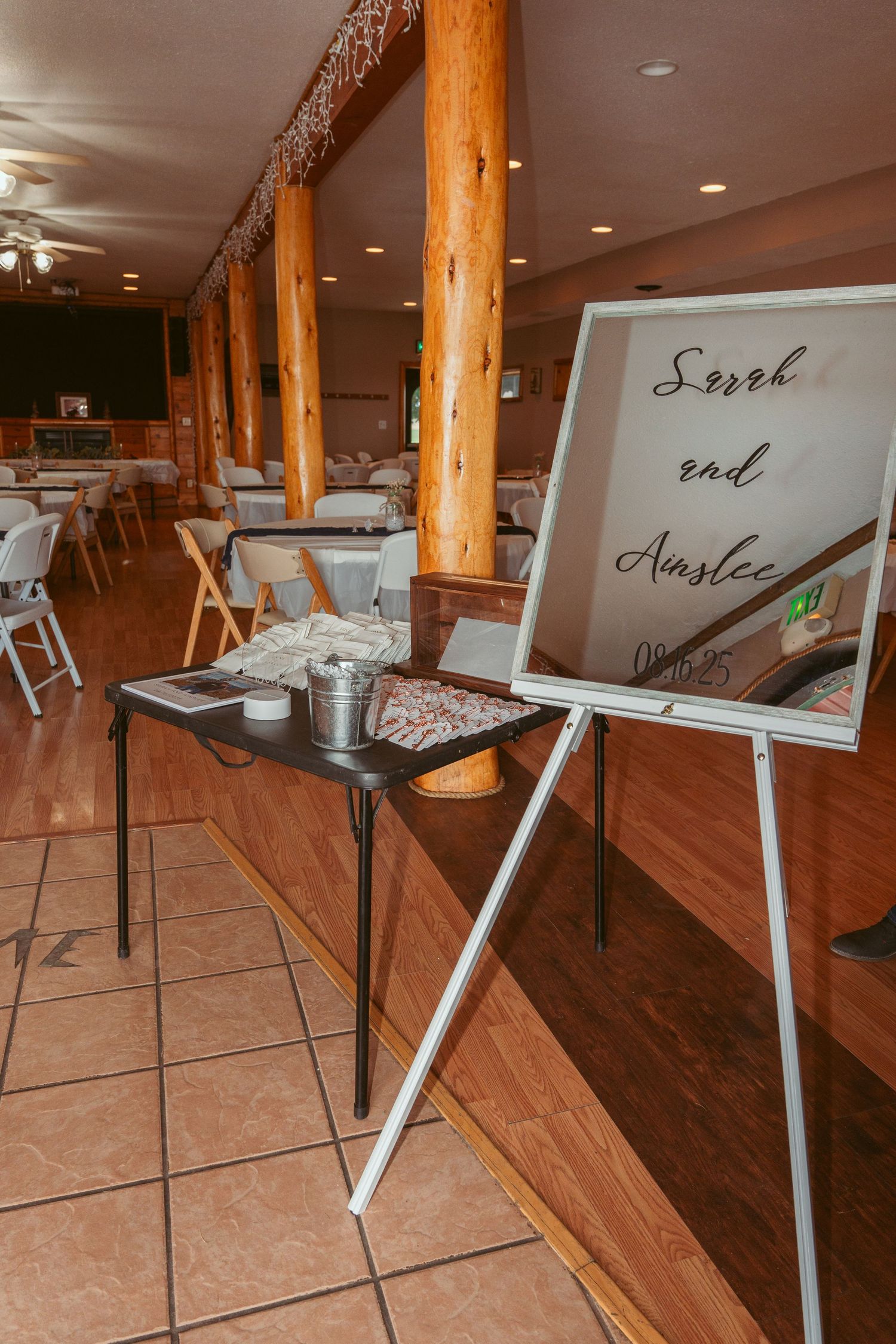 Interior of an event space with wooden columns, tile flooring and a welcome sign on an easel stand near dining tables.