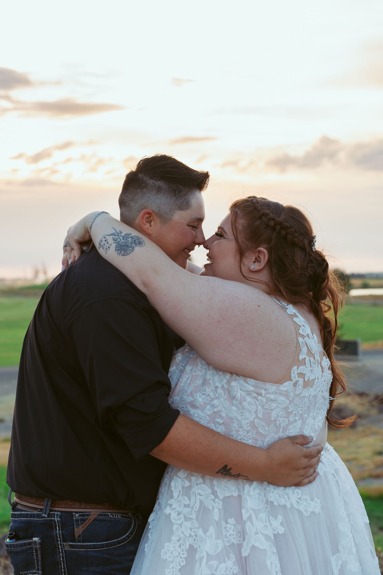 A couple shares a romantic kiss during sunset in a rural outdoor setting with rolling fields in the background.