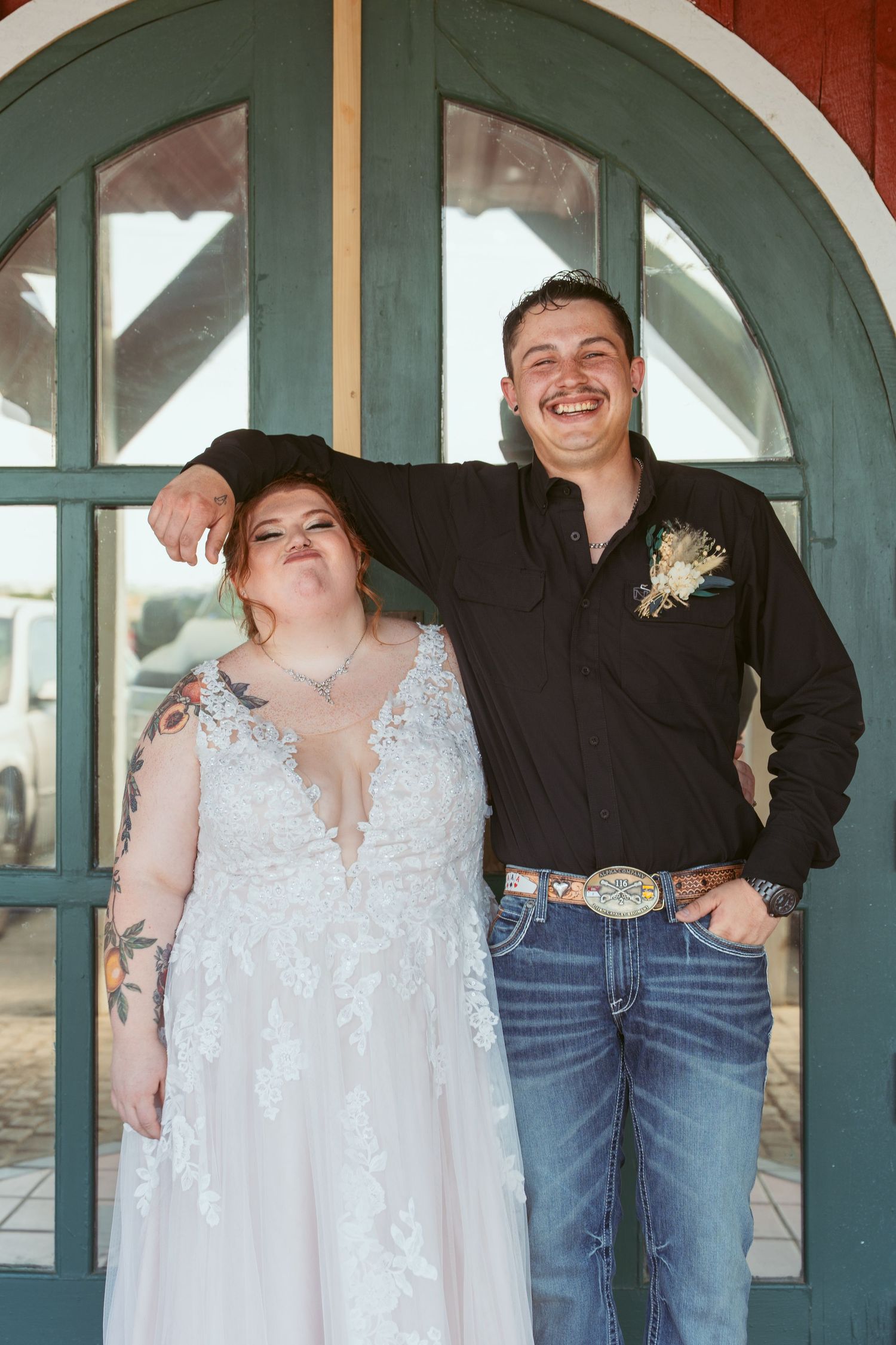 Wedding couple poses casually in front of arched teal door on red building.