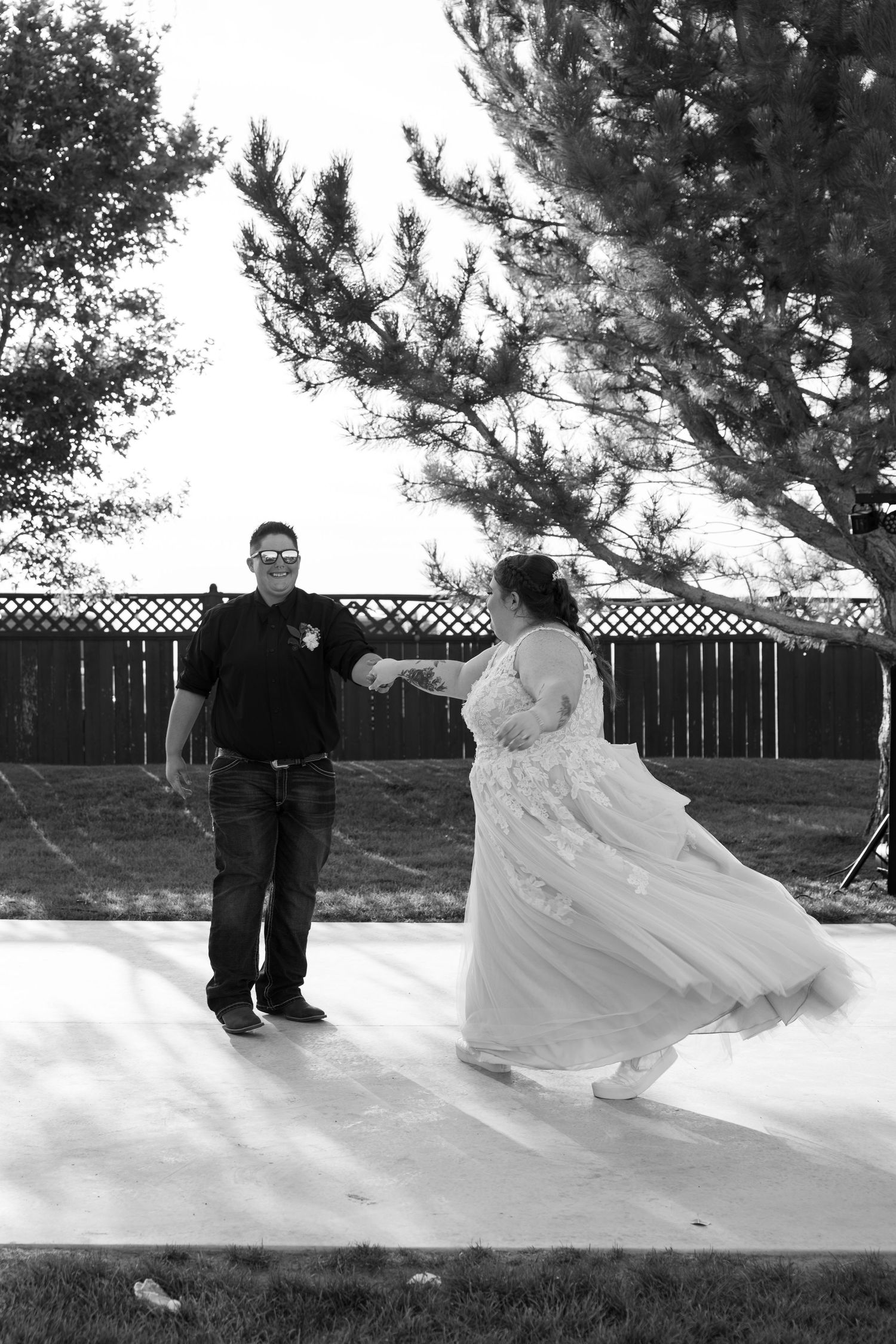 A couple dances together on an outdoor dance floor with pine trees and a wooden fence in the background.
