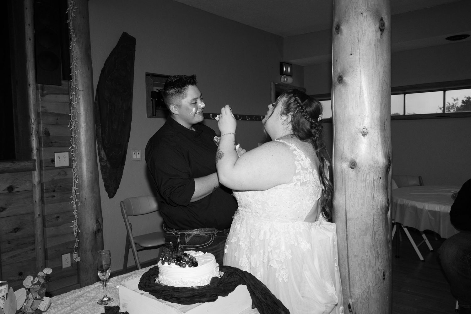 Black and white photo of a couple feeding each other wedding cake by a wooden pillar.