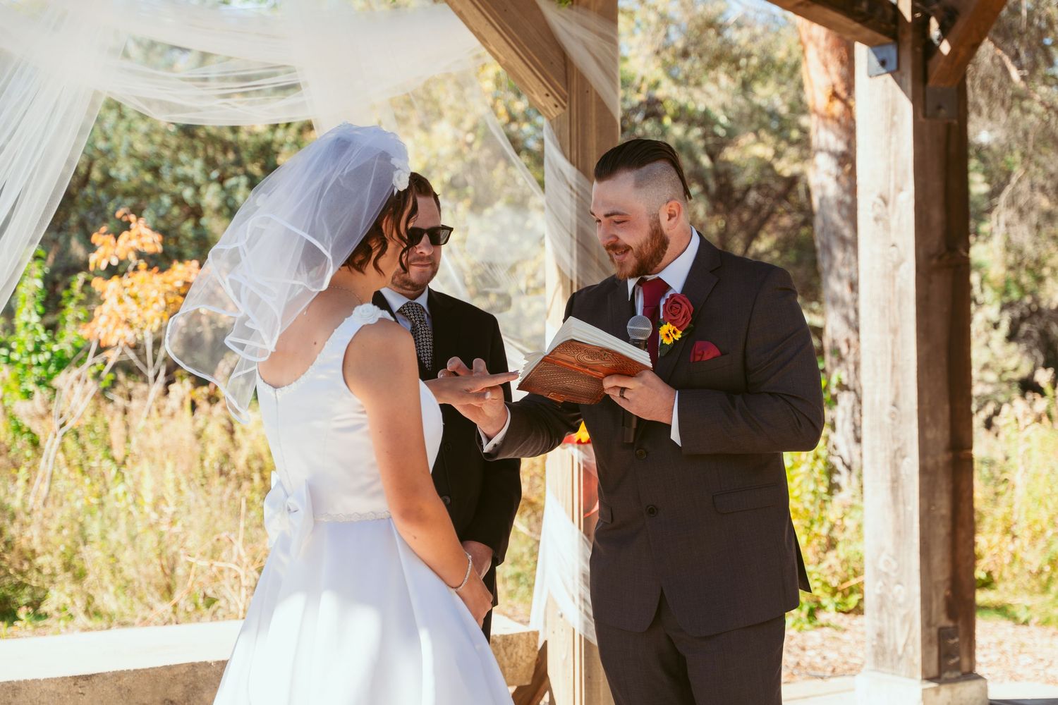 Wedding ceremony taking place under a rustic wooden pergola with white fabric draping.