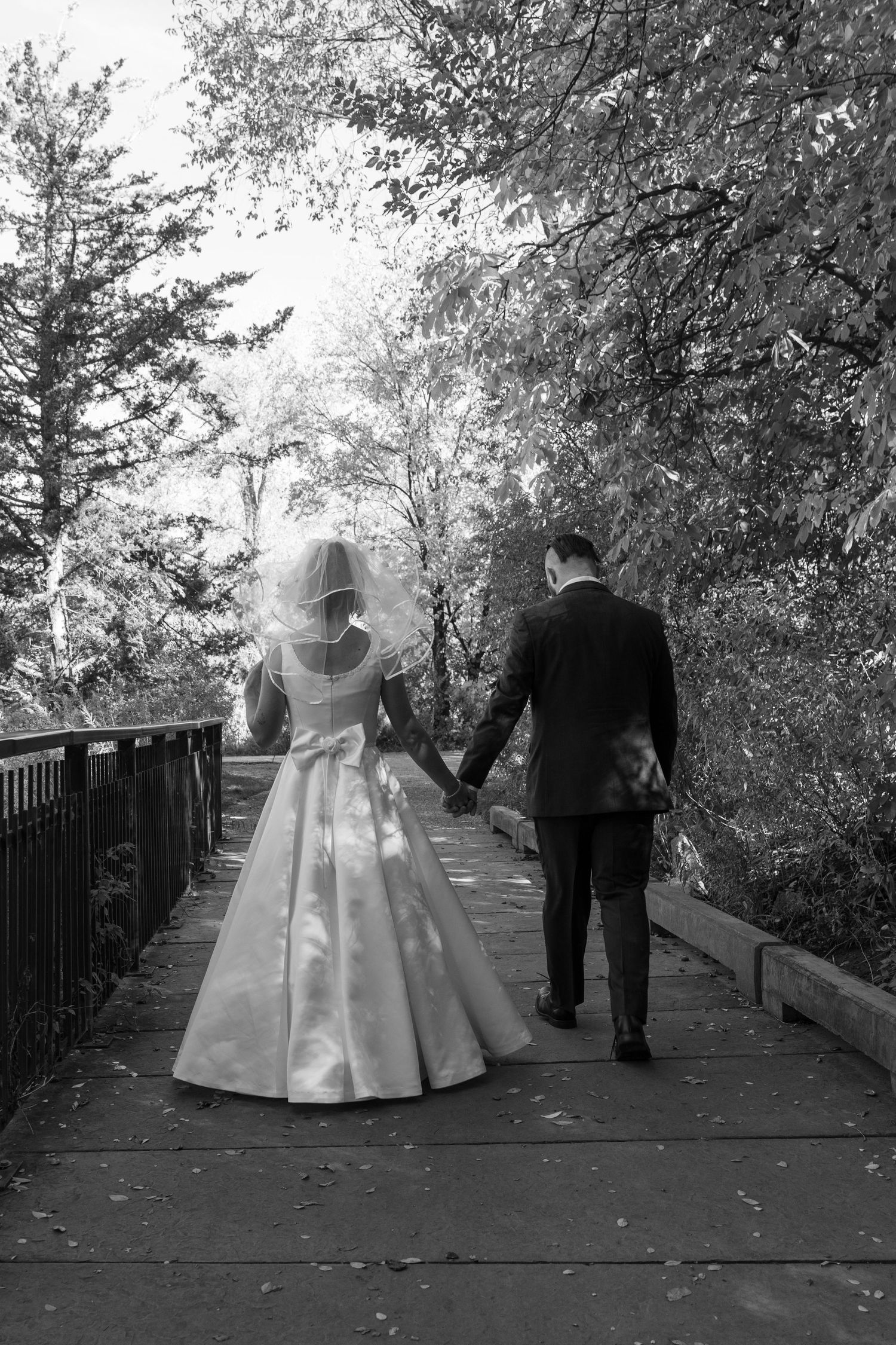 Black and white photo of newlyweds walking along elevated wooden pathway through trees.