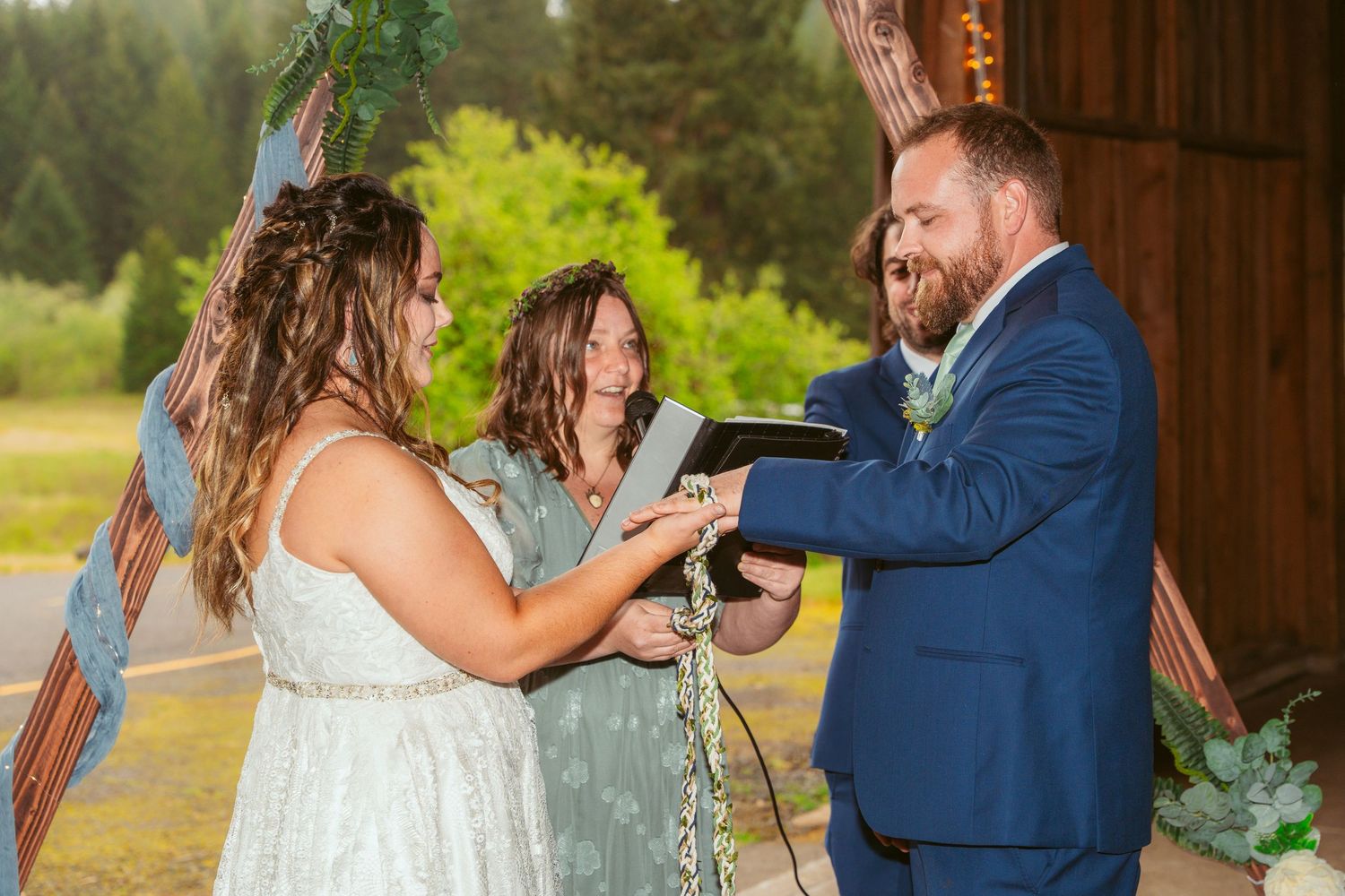 Wedding ceremony taking place in sunlit rustic barn with forest backdrop.
