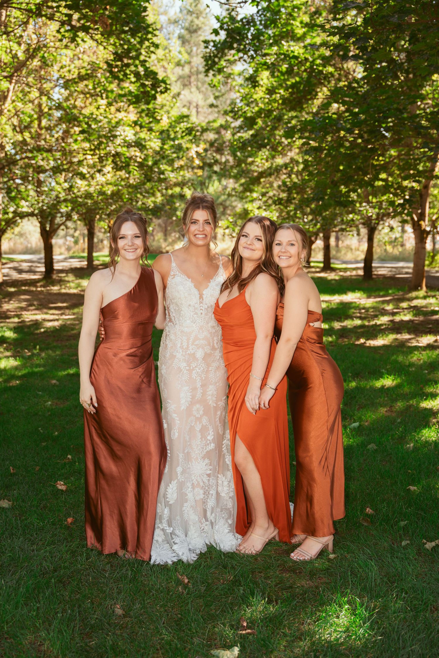 Wedding party in earth-toned dresses stands together in a wooded outdoor setting.
