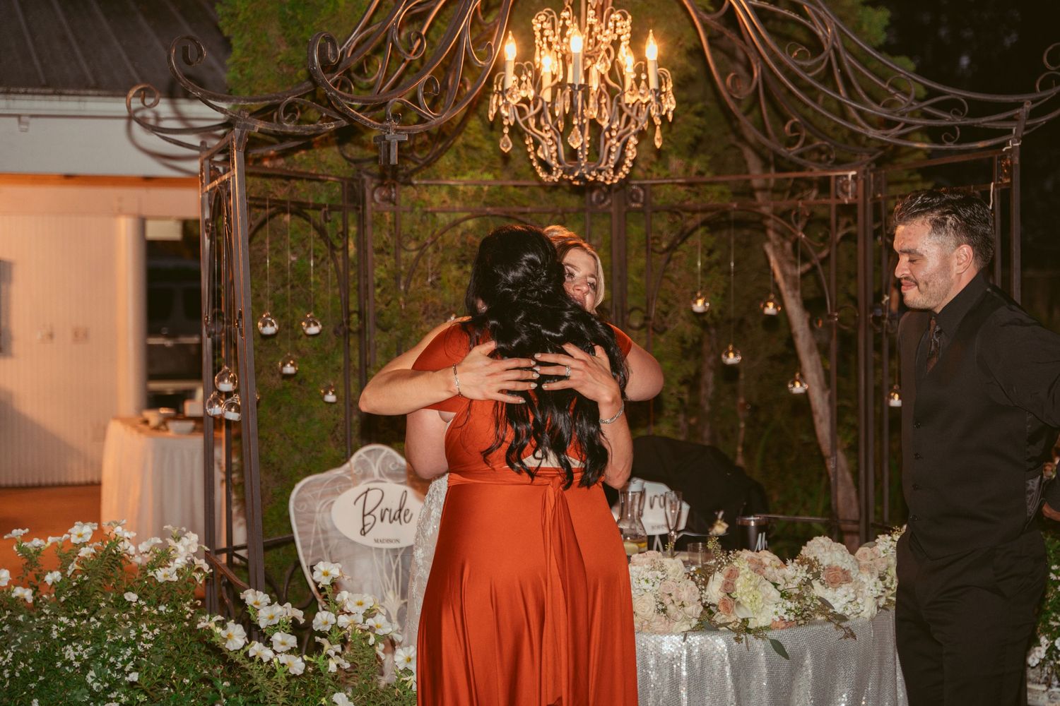 Wedding reception decor features elegant chandeliers and white floral arrangements on rustic wooden tables.